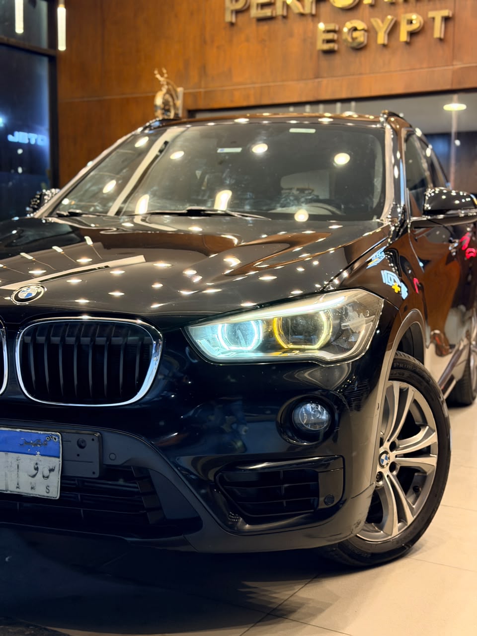 Black BMW X1 in a showroom, ceiling lights mirrored across the bonnet